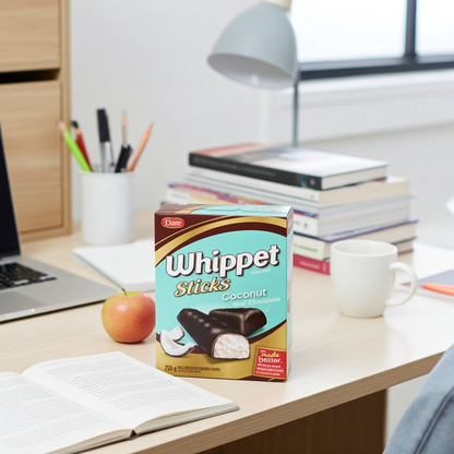 A box of Dannon Whippet Sticks Coconut ice cream sandwich sticks sits on a light wood desk in a bright, modern home office setting, surrounded by books, a laptop, an apple, and a mug.